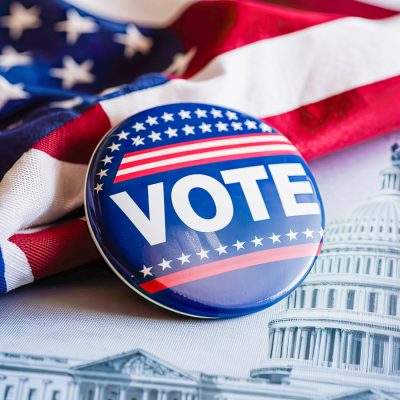 Round “VOTE” button with stars and stripes resting on an American flag, with the U.S. Capitol behind it