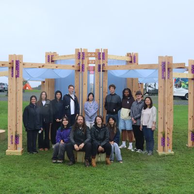 A group of 14 people pose together inside an open timber pavilion on a grassy field, with light blue fabric draped overhead and between the wooden posts.
