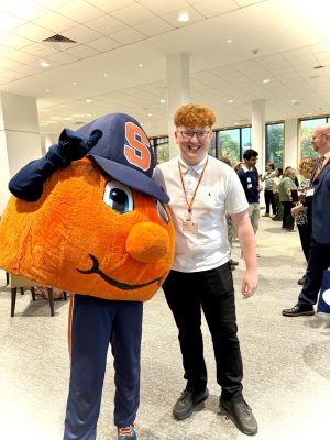 Smiling young man in a white polo poses with Otto the Orange, the Syracuse University mascot, at a reception.