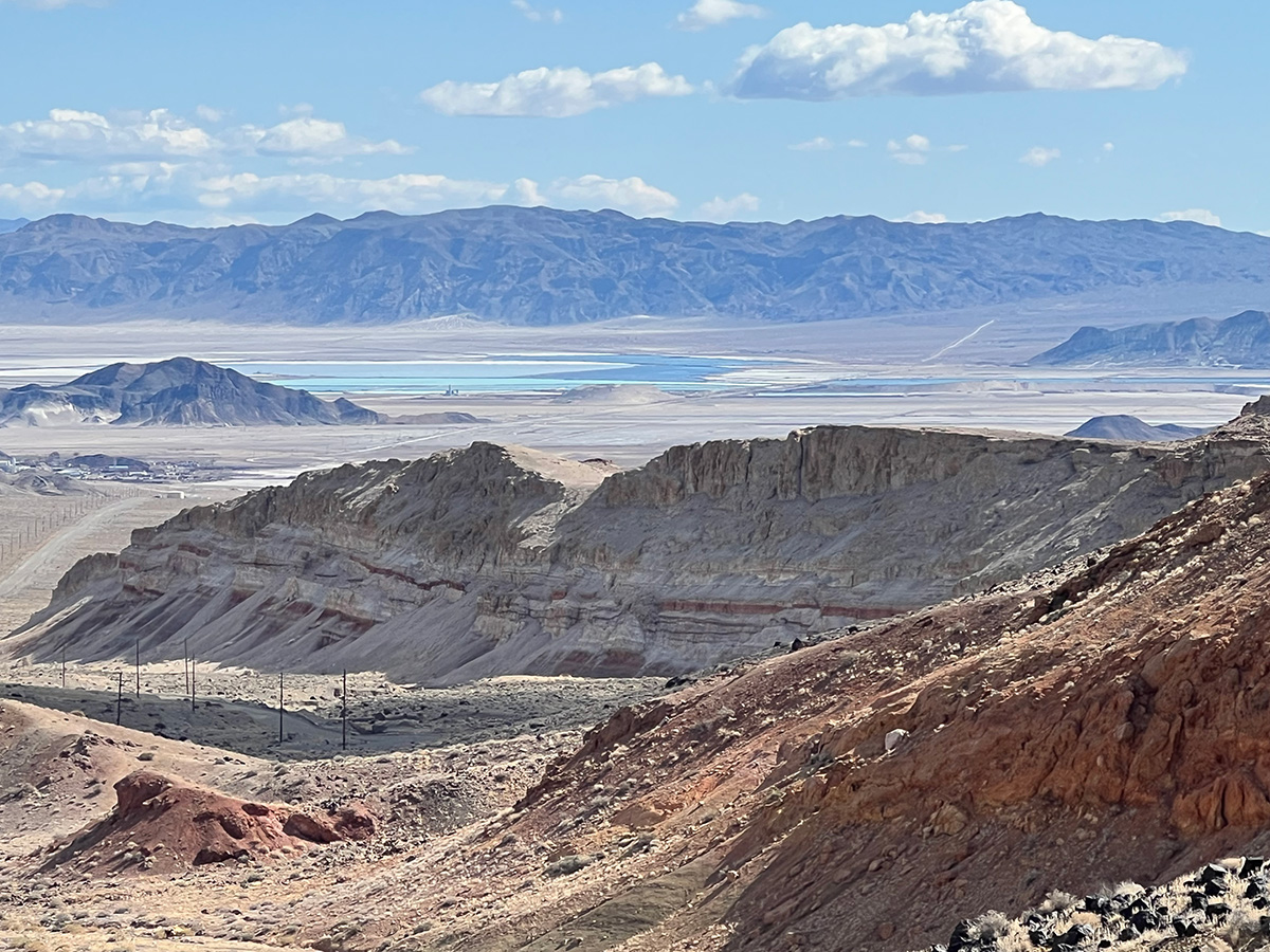 Desert valley with striped red and tan rock ridges, blue-green ponds on a distant basin, and mountains under a partly cloudy sky.
