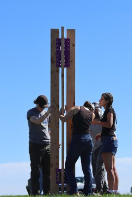 Four people stand around a tall wooden column made of three vertical timber posts joined by purple metal brackets, holding it steady against a clear blue sky.