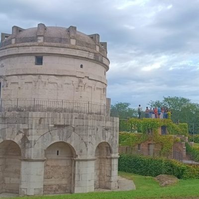Circular stone fortress tower with arched base, surrounded by greenery under a cloudy sky.