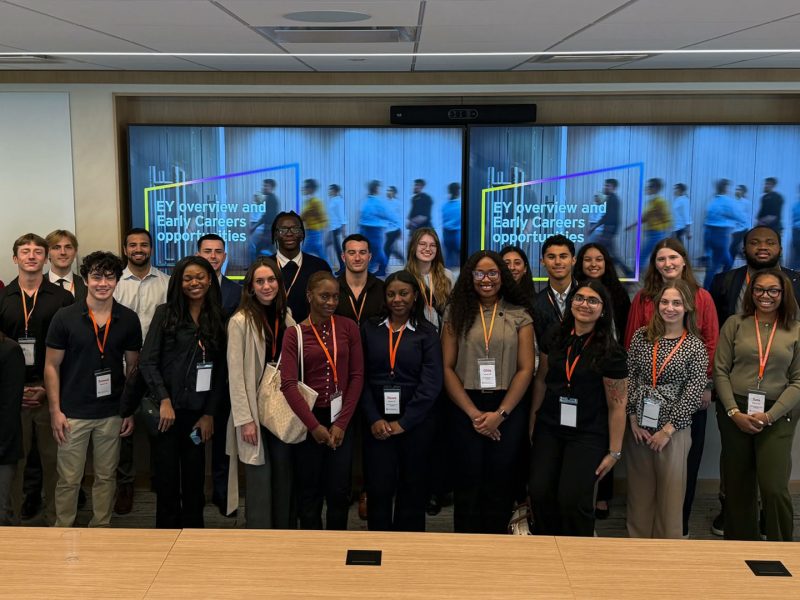 Students in lanyards pose in a conference room before screens reading “EY overview and Early Careers.”