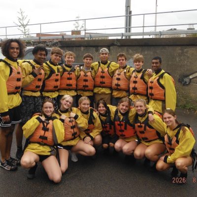 Sixteen students in jackets and life vests pose together as part of a rafting outing.