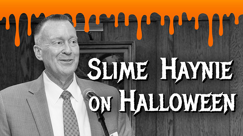 Black-and-white photo of a smiling man in a suit and tie at a microphone, with orange slime dripping across the top and text reading “Slime Haynie on Halloween”