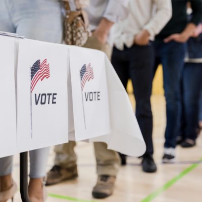 A line of voters stands in a gymnasium beside a table with signs reading “Vote” and featuring American flags.